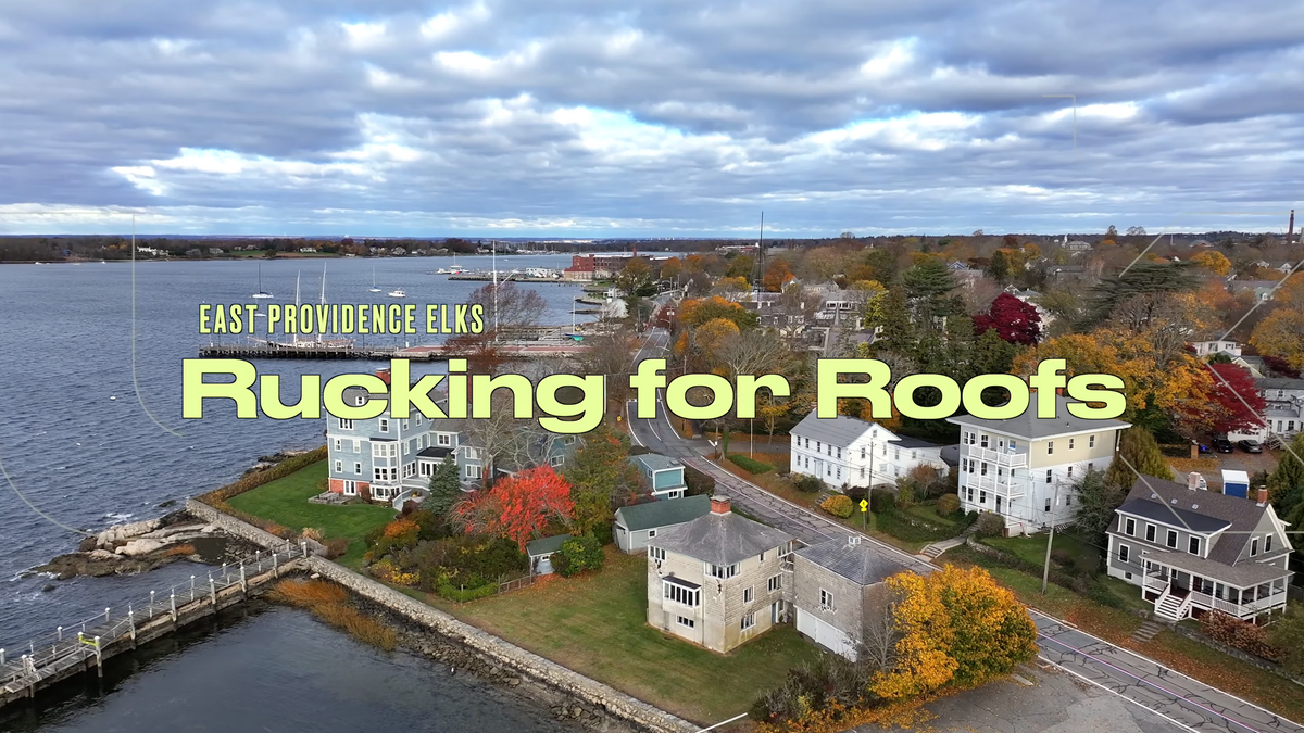 Aerial view of a waterfront community with houses and a marina, featuring text 'East Providence Elks Rucking for Roofs'.